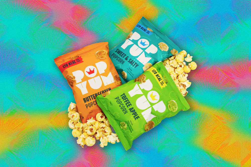 Colourful vegan & gluten-free popcorn packets on a psychedelic background, pictured with a pile of popcorn.