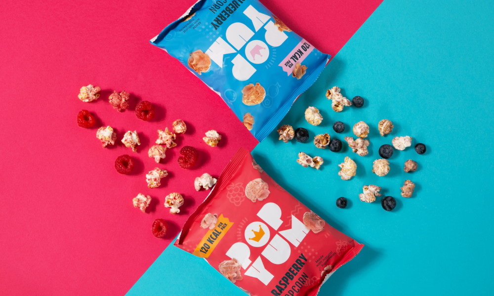 Colourful Pop Yum Veganuary popcorn packets, pictured on a pink and blue gradient beside popcorn piles.