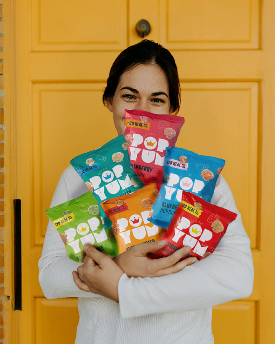 A happy woman holding colourful Pop Yum popcorn packets in front of a yellow door. 