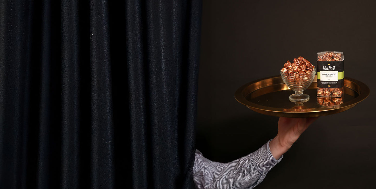 Hand popping out of a curtain holding a gold tray with Gourmet Moments vegan Belgian chocolate-drizzled popcorn.