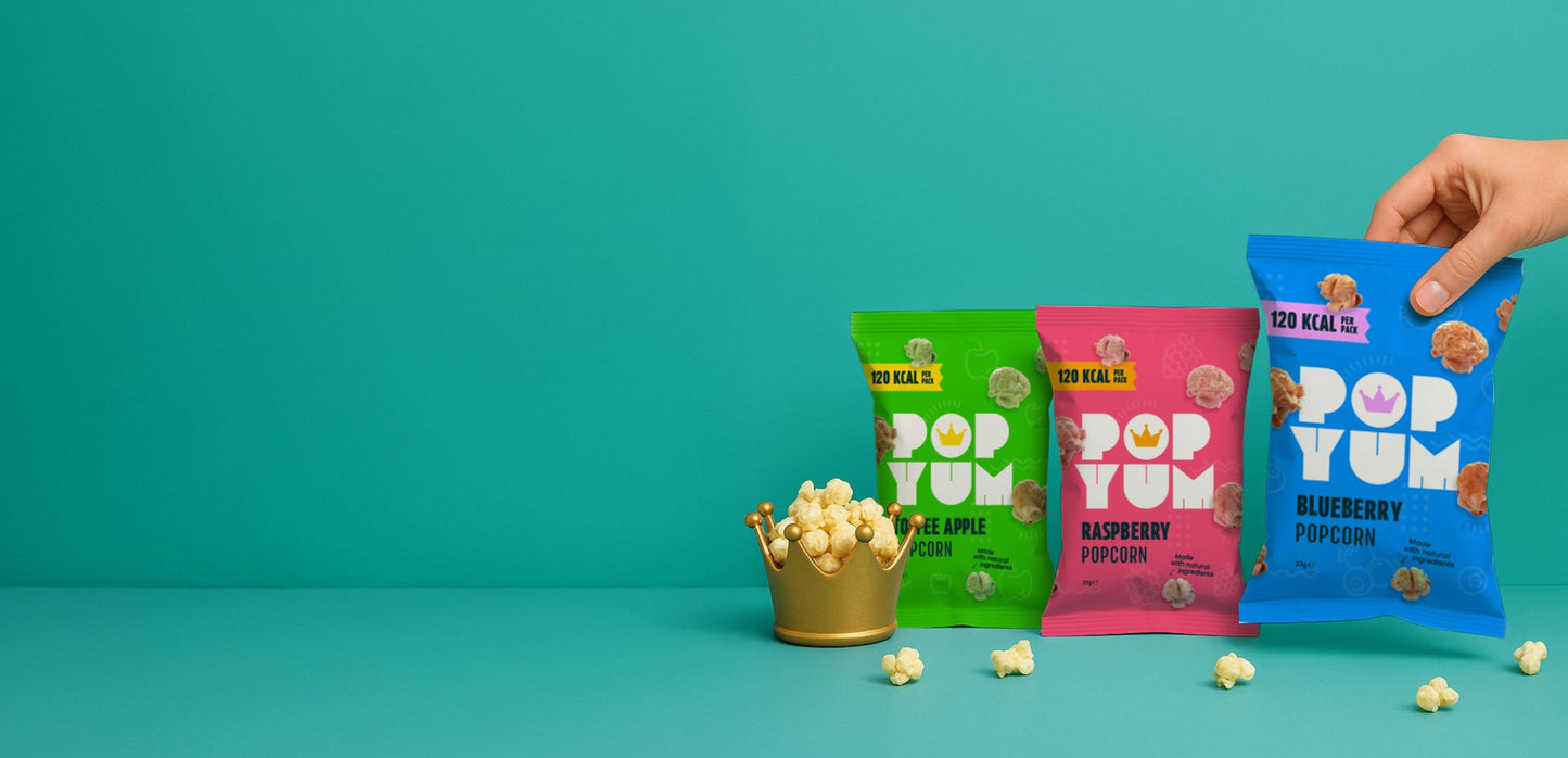 Three colourful Pop Yum popcorn packets on a teal background with scattered popcorn kernels & a crown.