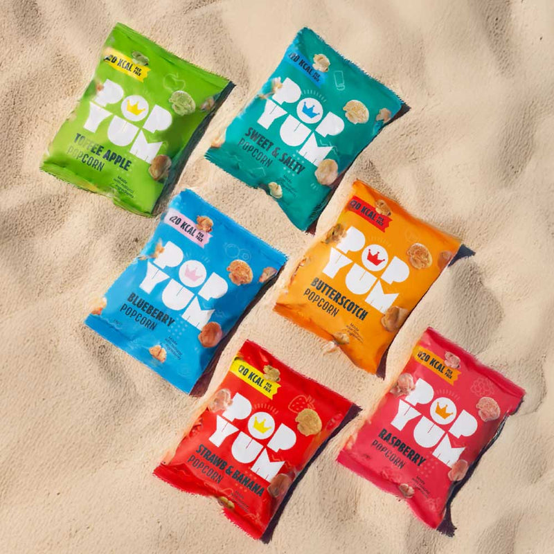 Sweet & salty, butterscotch, raspberry, toffee apple, strawberry banana, and blueberry popcorn packets on a sandy surface.