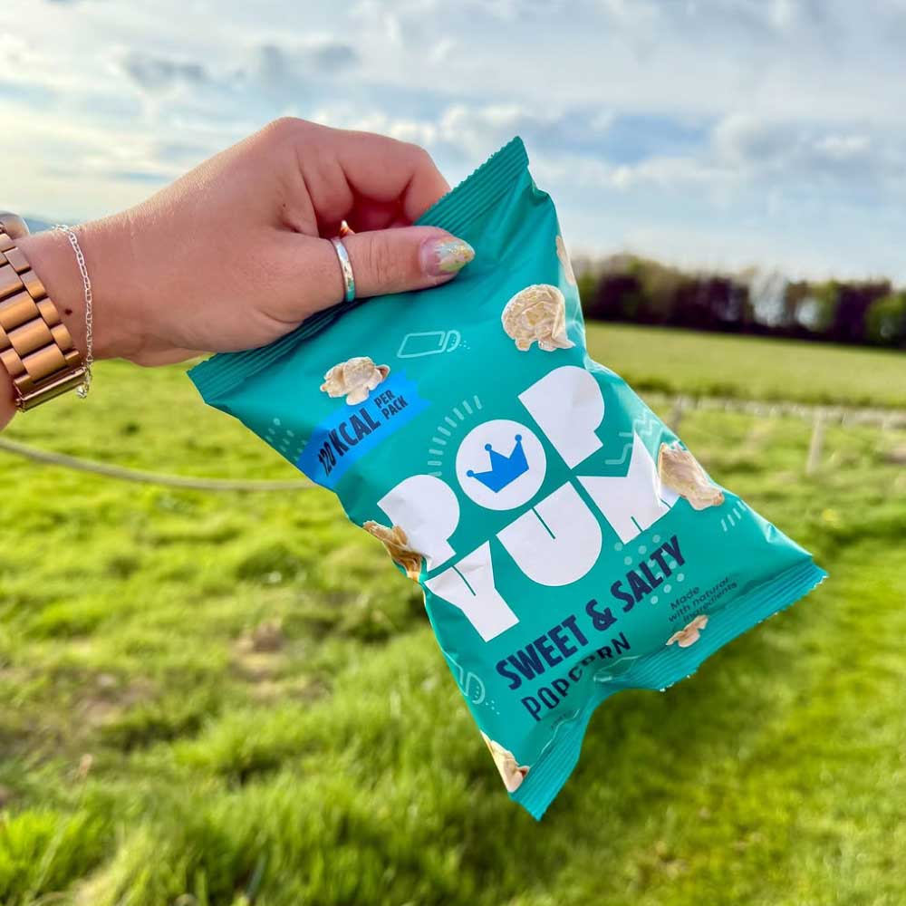 Hand holding a teal Pop Yum sweet & salty-flavoured gluten-free popcorn packet against a green field background.