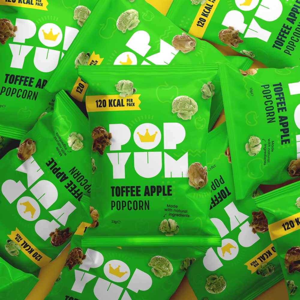 Multiple green Pop Yum toffee apple-flavoured vegan sweet popcorn packets stacked together on a yellow background.