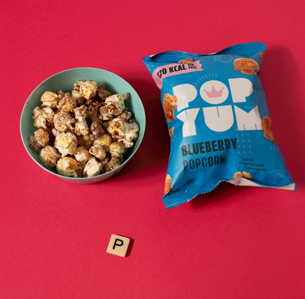 Animated image of Scrabble pieces, which spell out "Pop Yum", accompanied by a packet of Pop Yum Blueberry popcorn and a bowl.