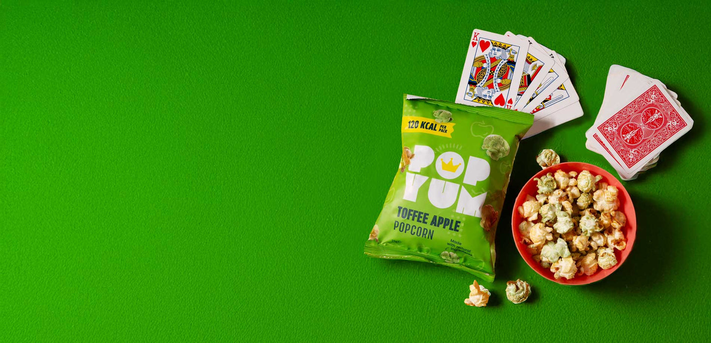 Pop Yum Toffee Apple flavoured green popcorn bag, a red bowl of popcorn, and playing cards on a green background.
