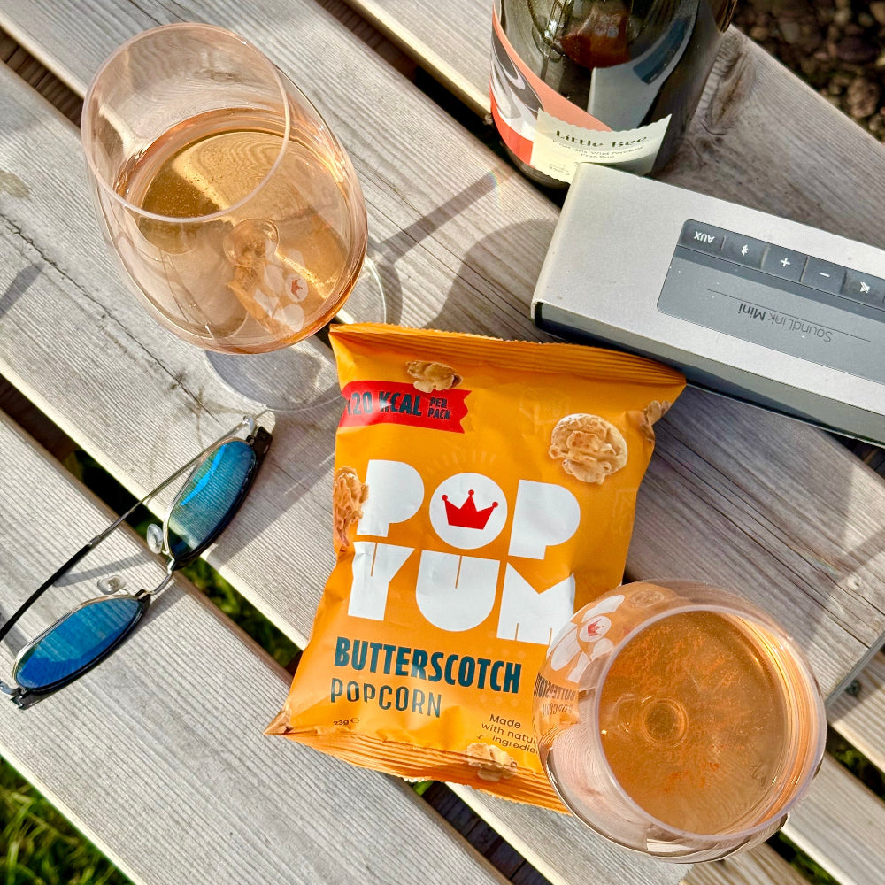 A pack of butterscotch popcorn is pictured on a wooden surface with a speaker, sunglasses, two wine glasses, and a bottle. 